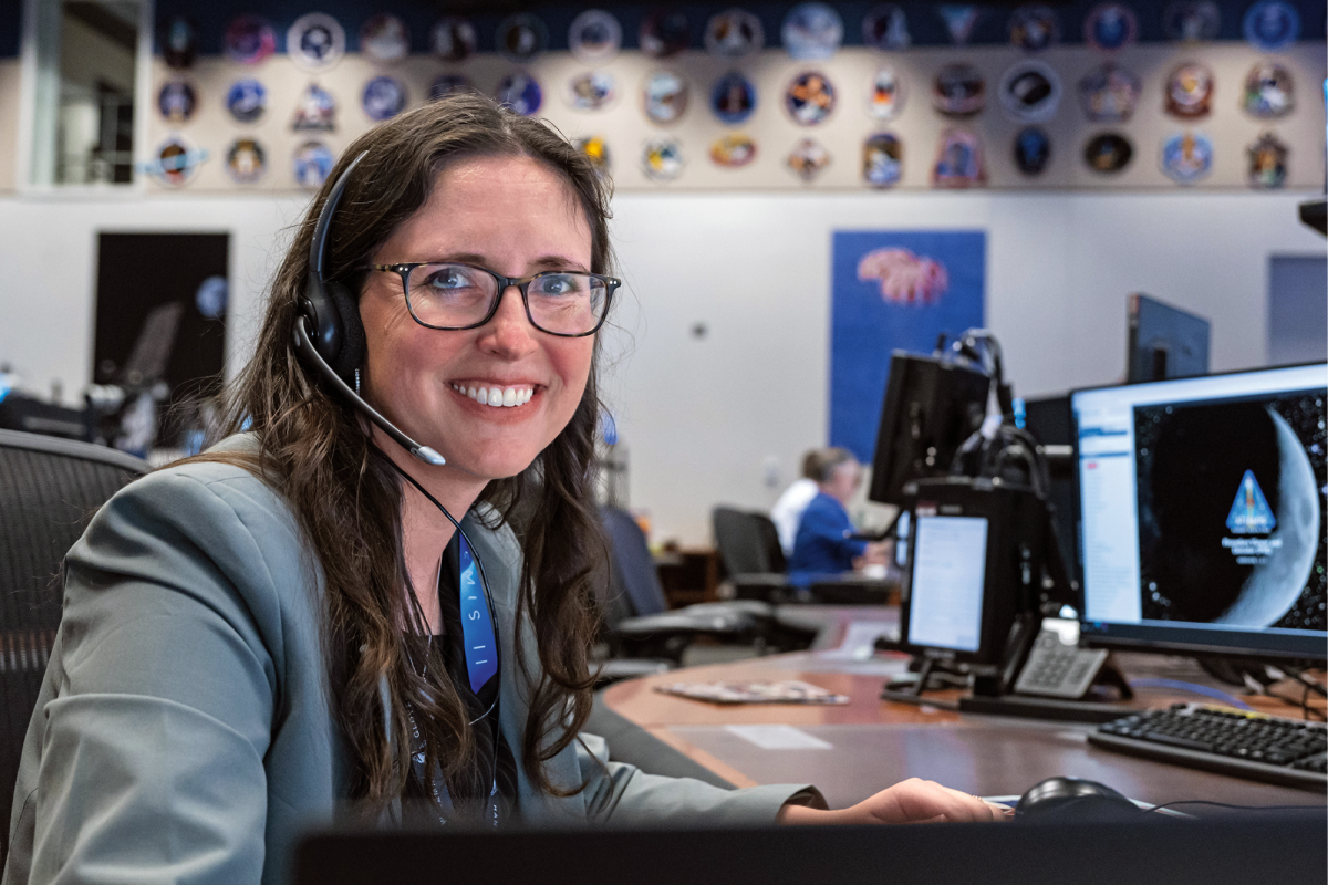 A portrait of a woman sitting at a desk in a NASA command center smiling at the camera